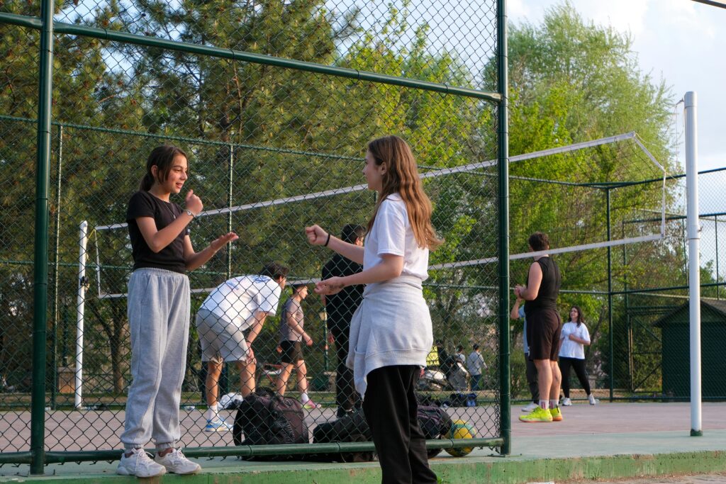 A group of teenagers engaged in a casual volleyball game on an outdoor court. Trees in the background.