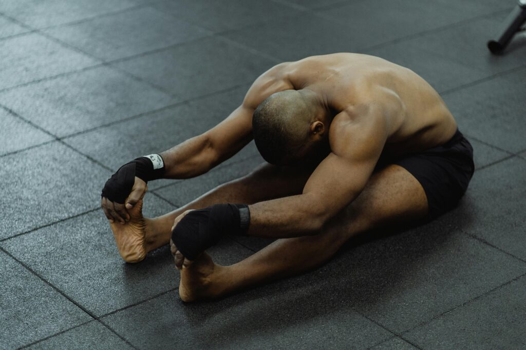 A shirtless man stretches on a gym floor, barefoot and wearing black shorts, focused on flexibility.